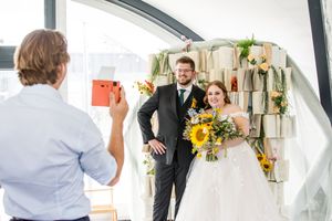 A wedding photographer captures a couple posing in front of a decorative book wall backdrop with sunflowers.