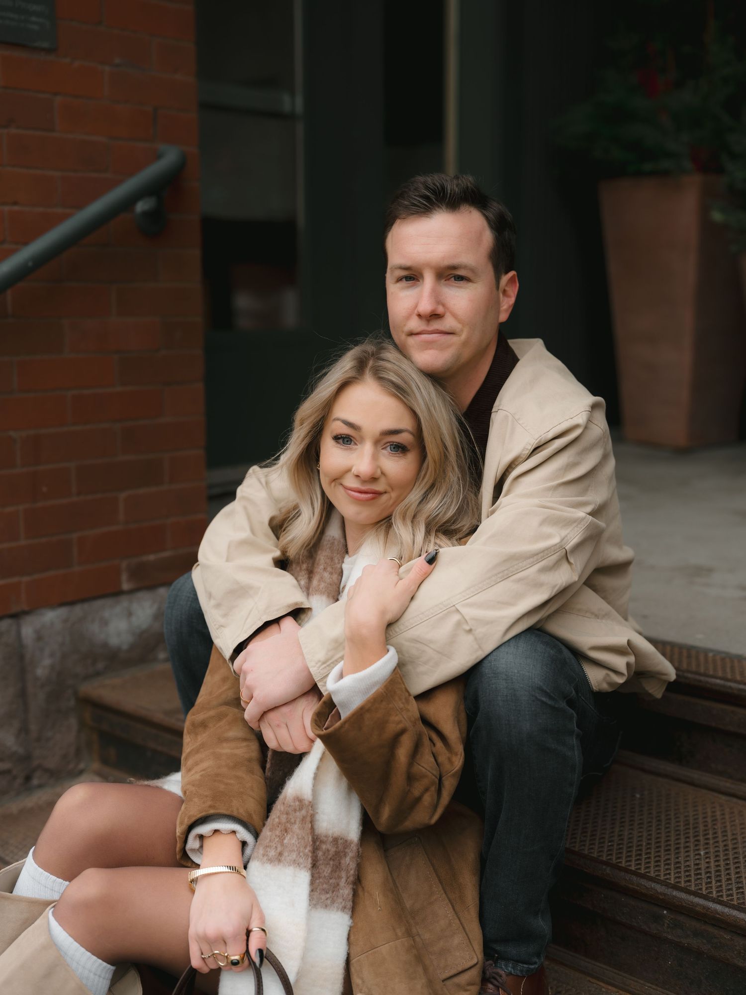 A couple in casual attire sits together on brick steps sharing intimate moments in a romantic photoshoot.