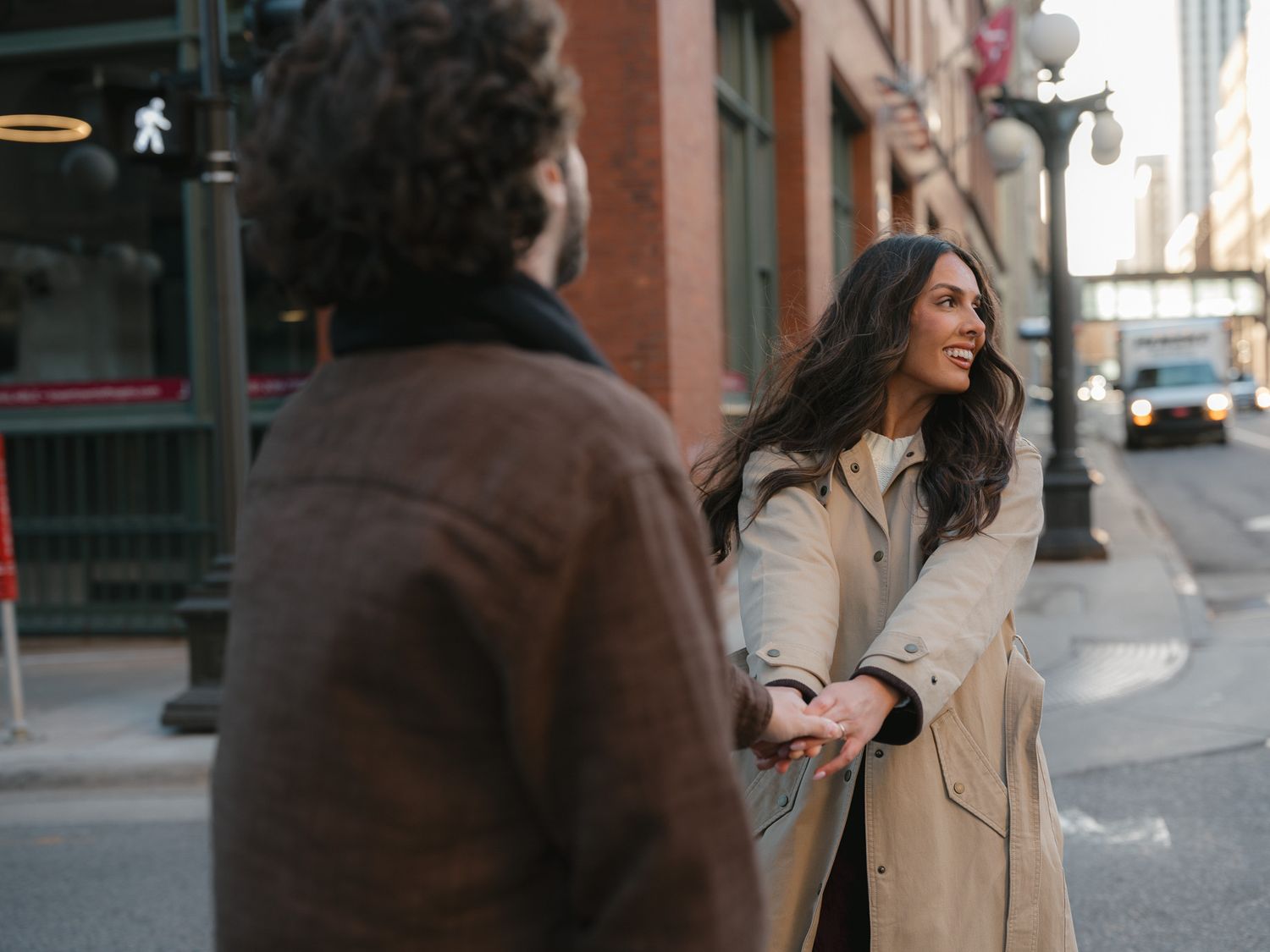 Woman in beige coat walks down urban street while looking over her shoulder on cloudy day.