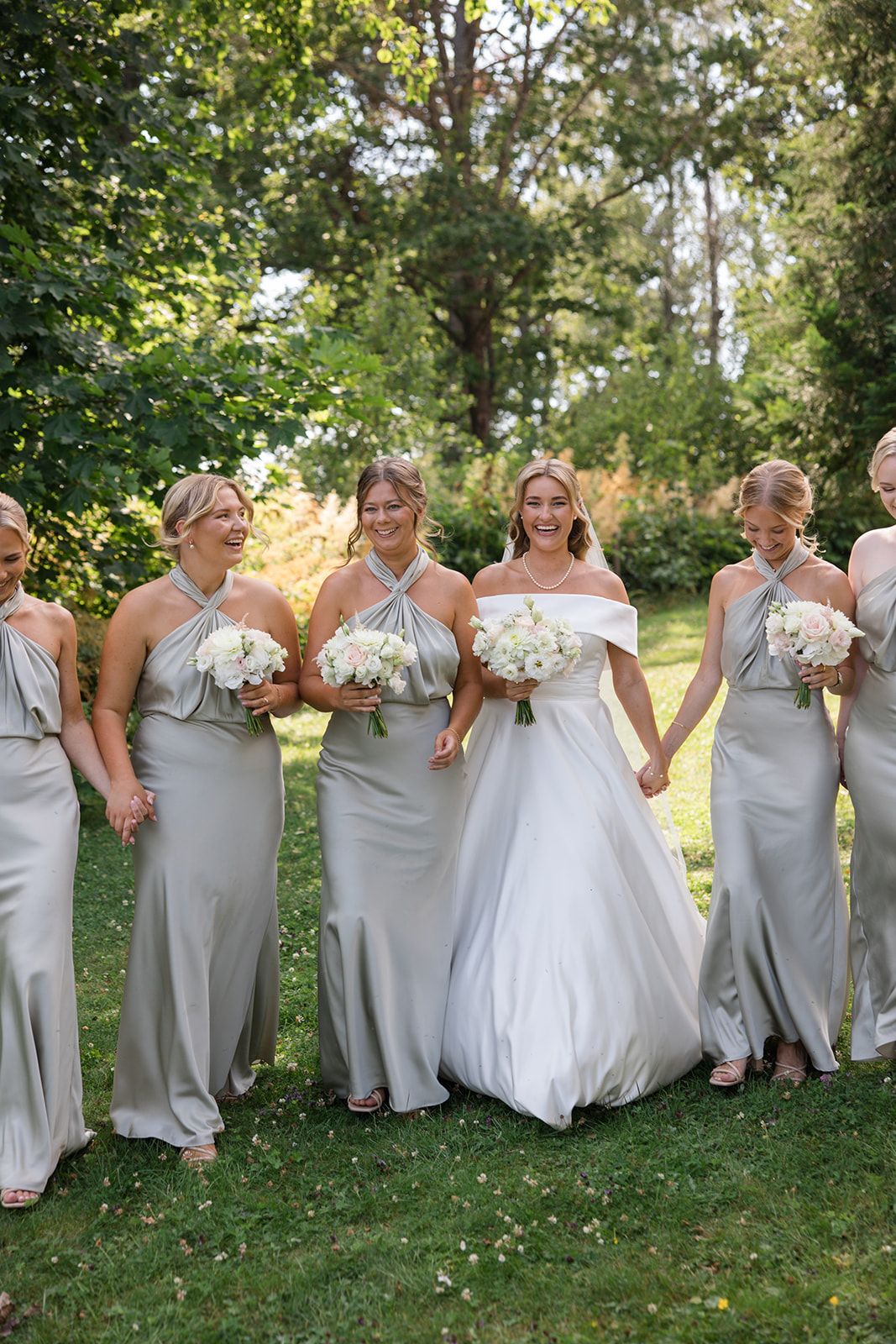 Wedding party in light gray dresses and white gown pose playfully on grassy hill during outdoor summer celebration.