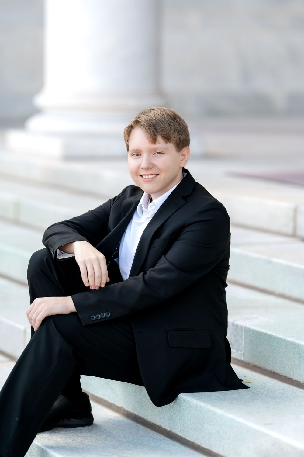 Smiling teenage boy in black suit sitting relaxed on marble steps of a classical building.