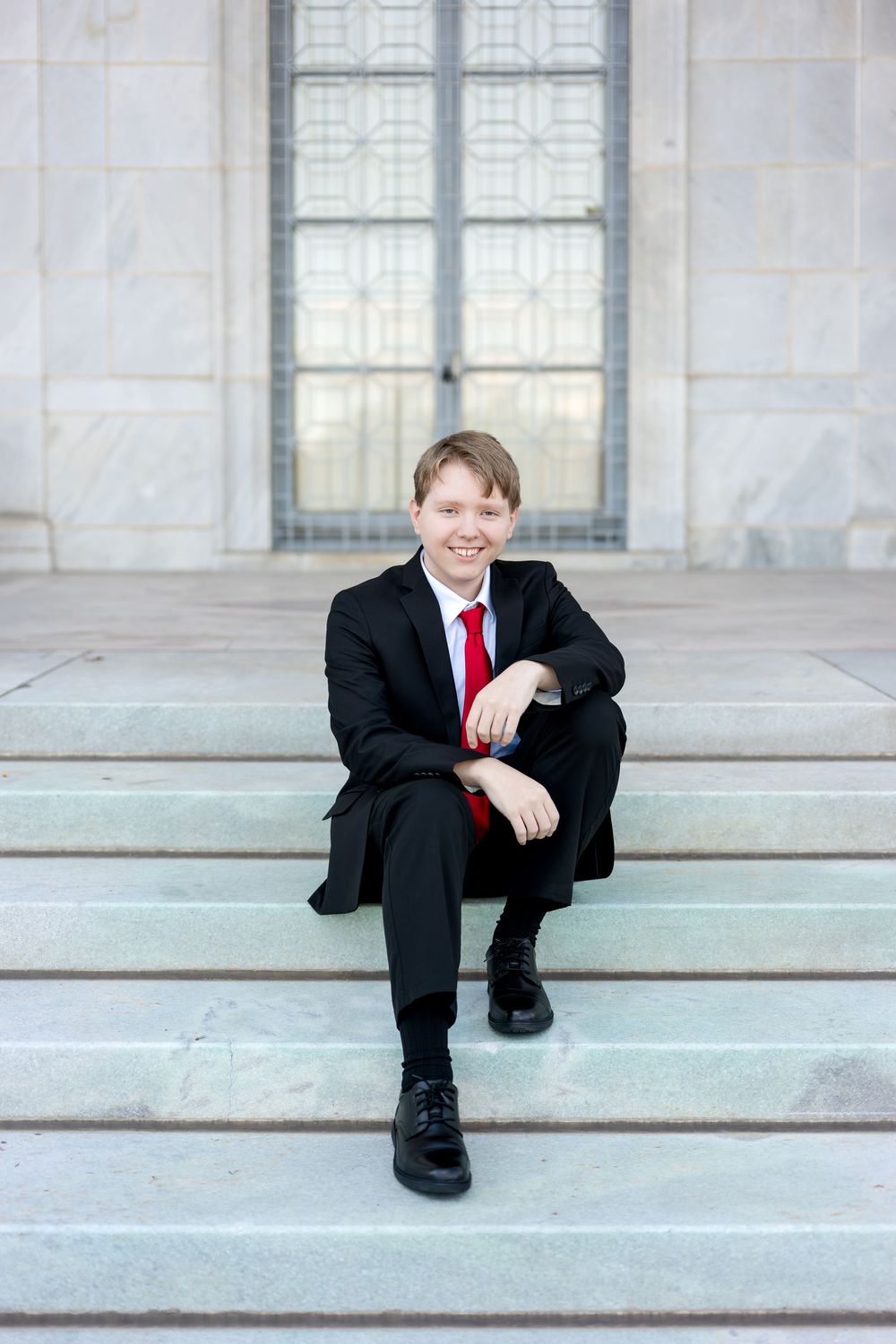Smiling boy in black suit and red tie sitting on stone steps outdoors.