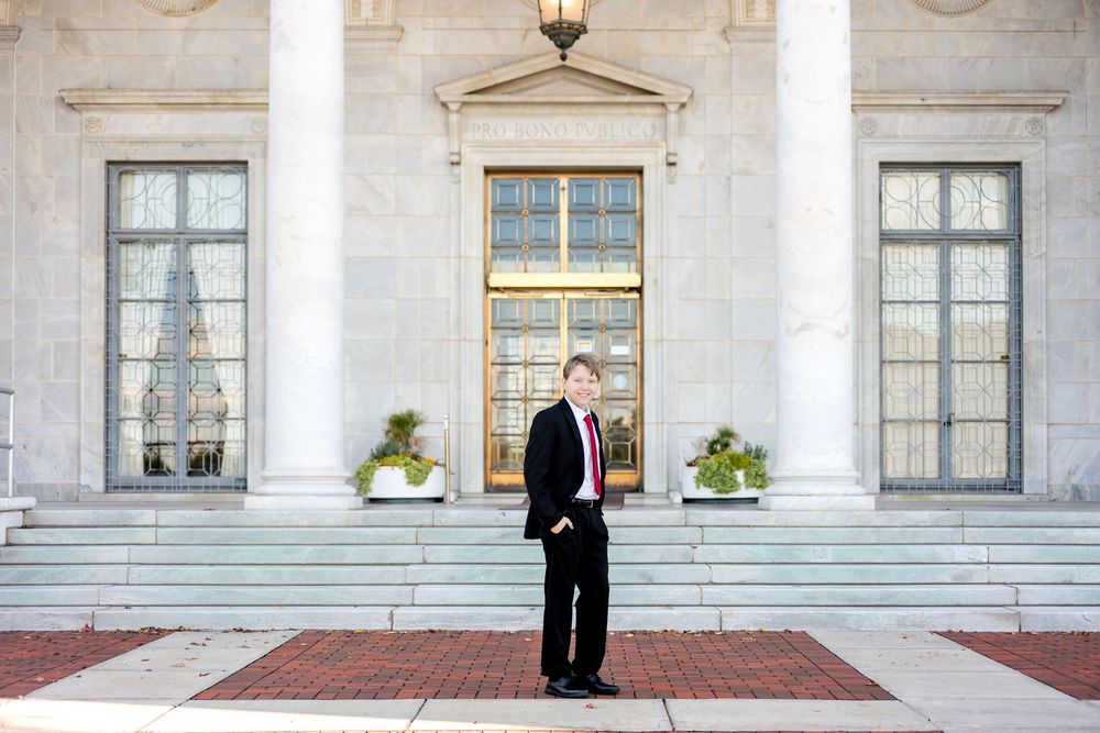 Young man in black suit and red tie standing before a grand neoclassical building.