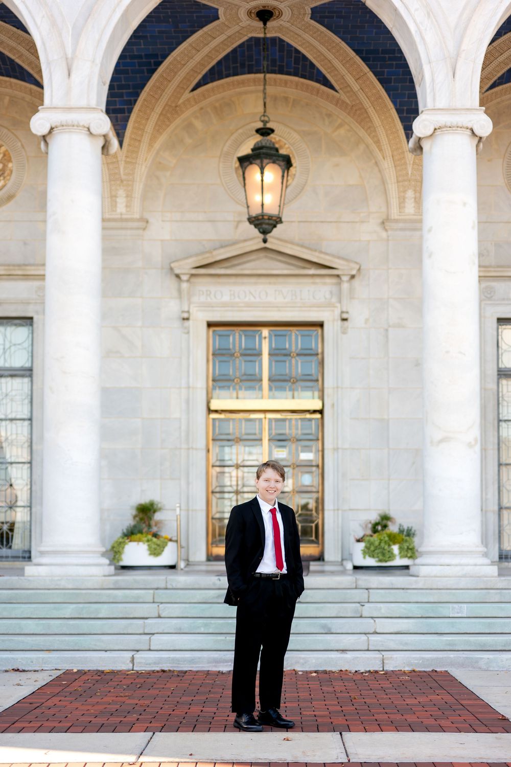 Smiling young man in black suit standing before a grand neoclassical building entrance.