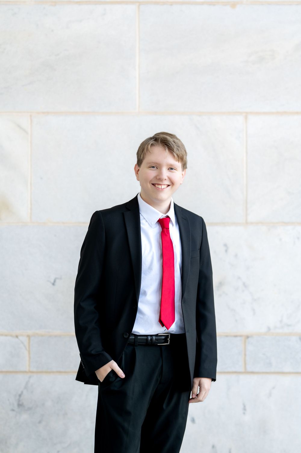 Smiling young man in black suit and red tie standing against a marble wall.