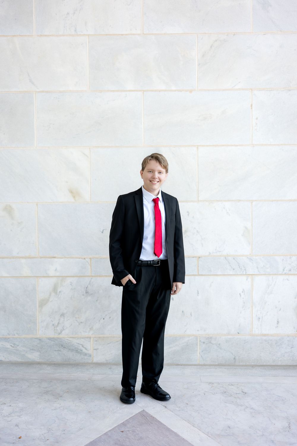 Young man in black suit and red tie standing against a marble wall, smiling.