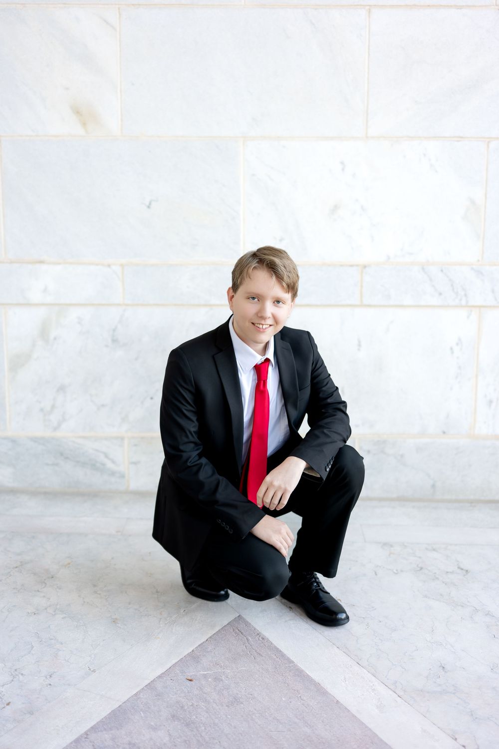 Young man in black suit and red tie crouching on marble floor, smiling at camera.