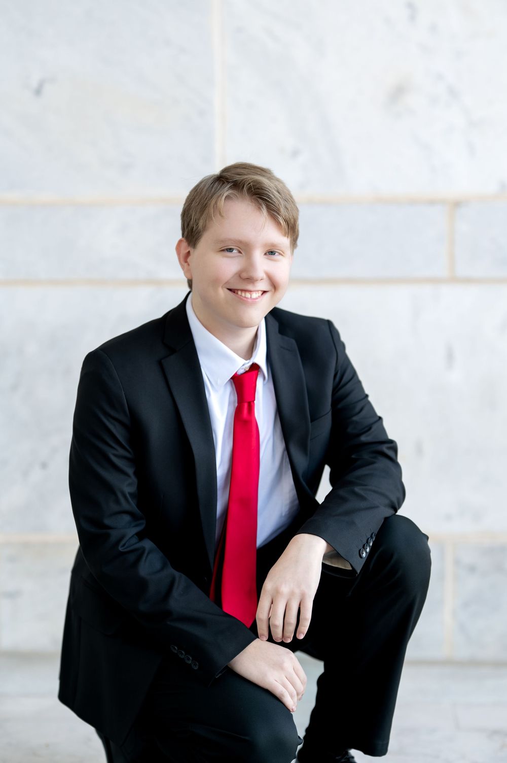 Smiling young man in black suit and red tie sitting against marble wall background.