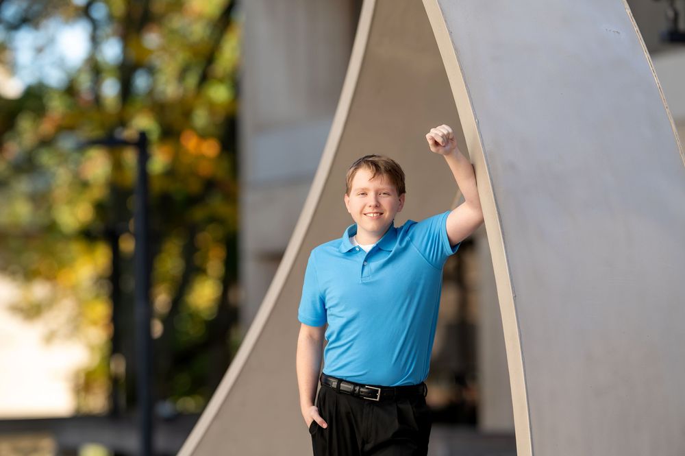 Young man in blue polo shirt raising fist triumphantly beside large metal arch sculpture.