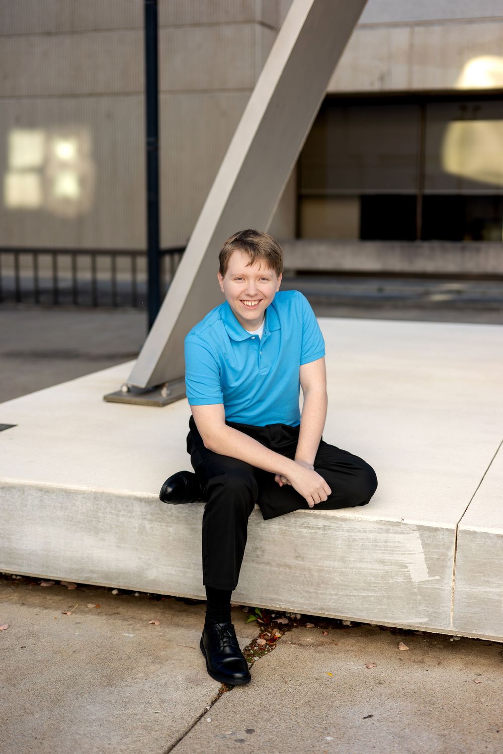 Smiling teenage boy in blue polo shirt sitting on concrete bench outside modern building.