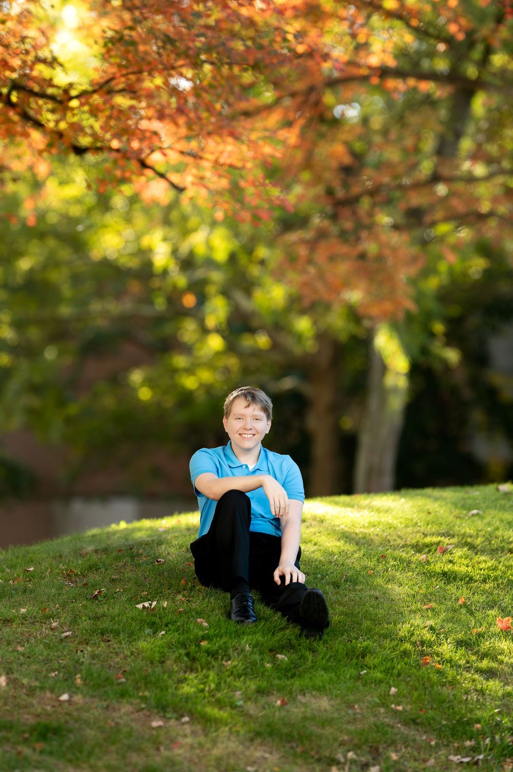 Smiling teenage boy in blue polo sitting cross-legged on green grass with autumn trees behind.