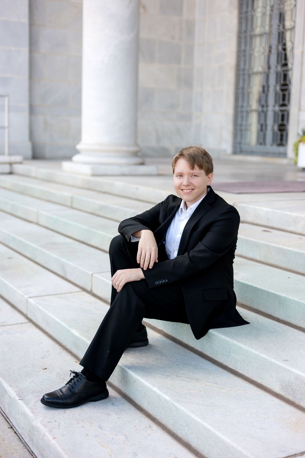 Smiling teenage boy in black suit sitting on wide marble steps of a grand classical building.