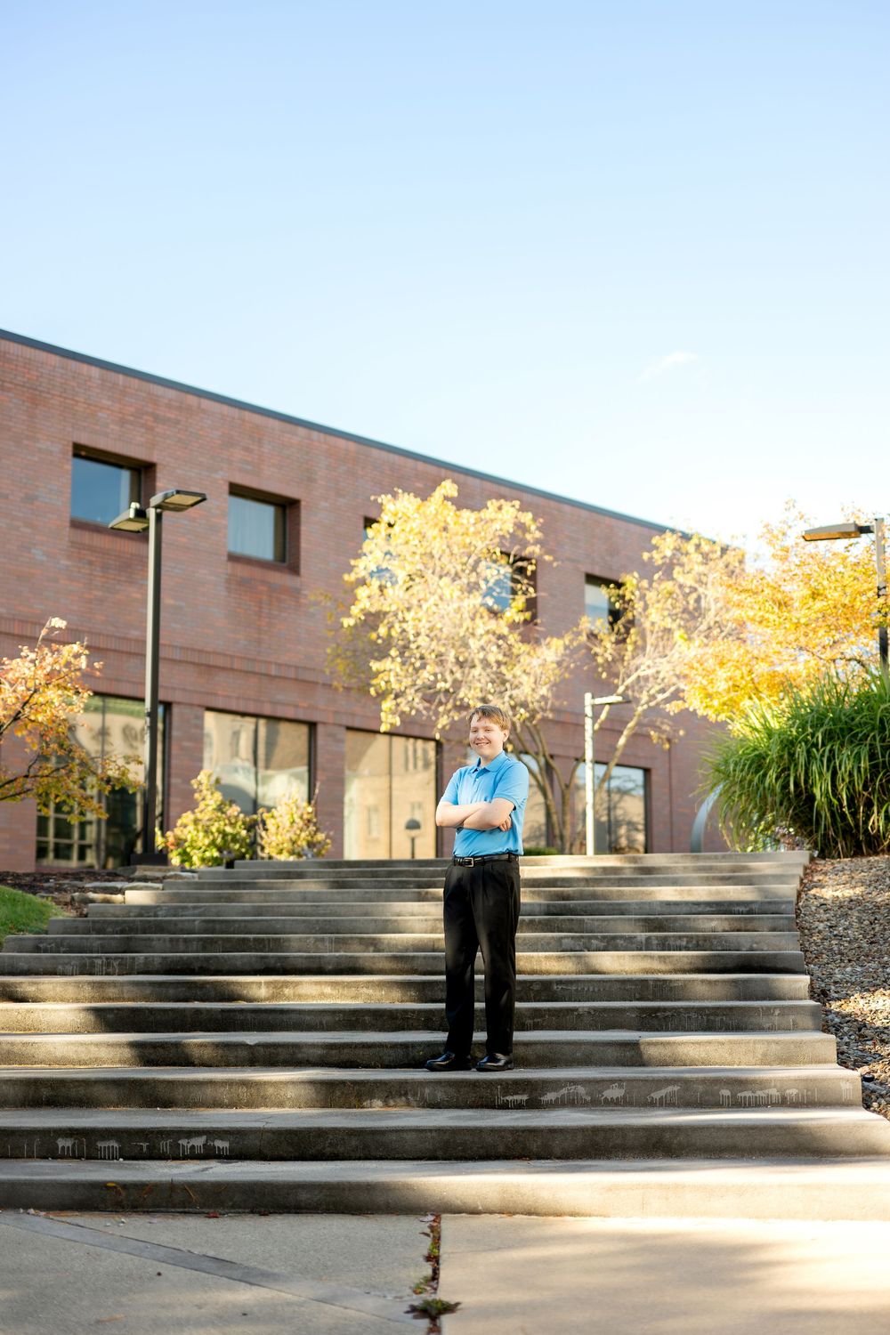 Teen boy in blue polo with arms crossed standing on steps outside brick building.