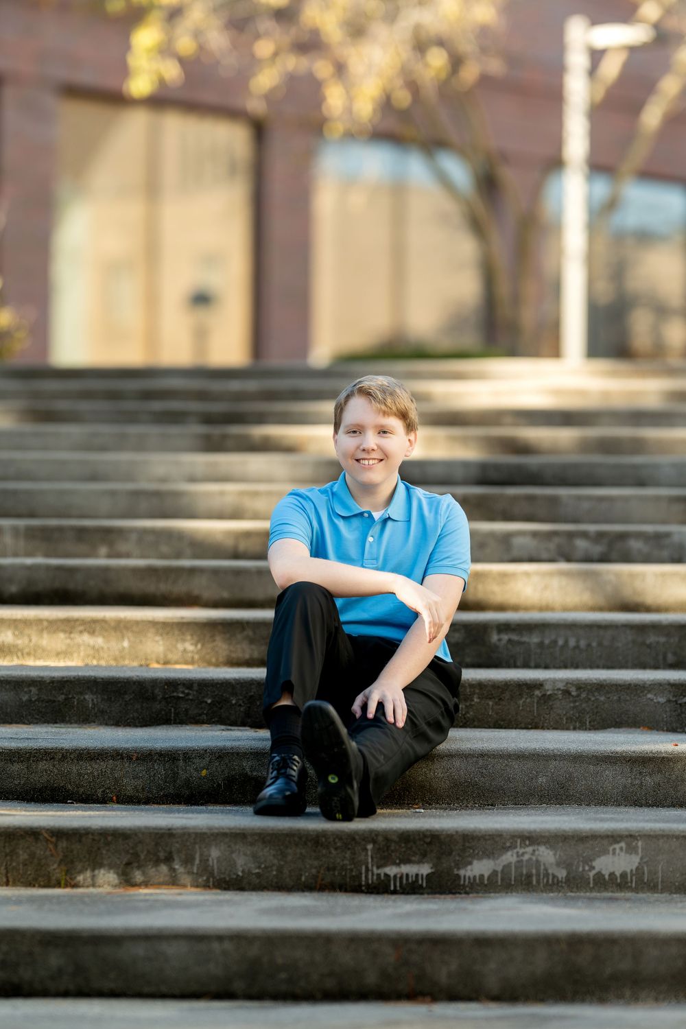 Happy teen boy in blue polo shirt sitting relaxed on outdoor concrete steps smiling.