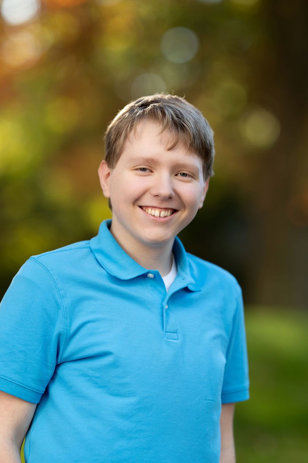 Close-up portrait of smiling teenage boy in blue polo shirt with green bokeh background.
