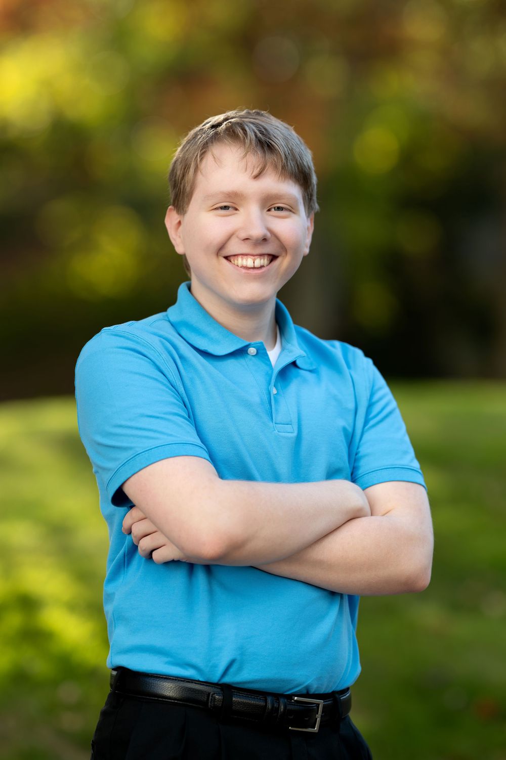 Confident smiling teen boy in blue polo shirt with arms crossed in autumn park.