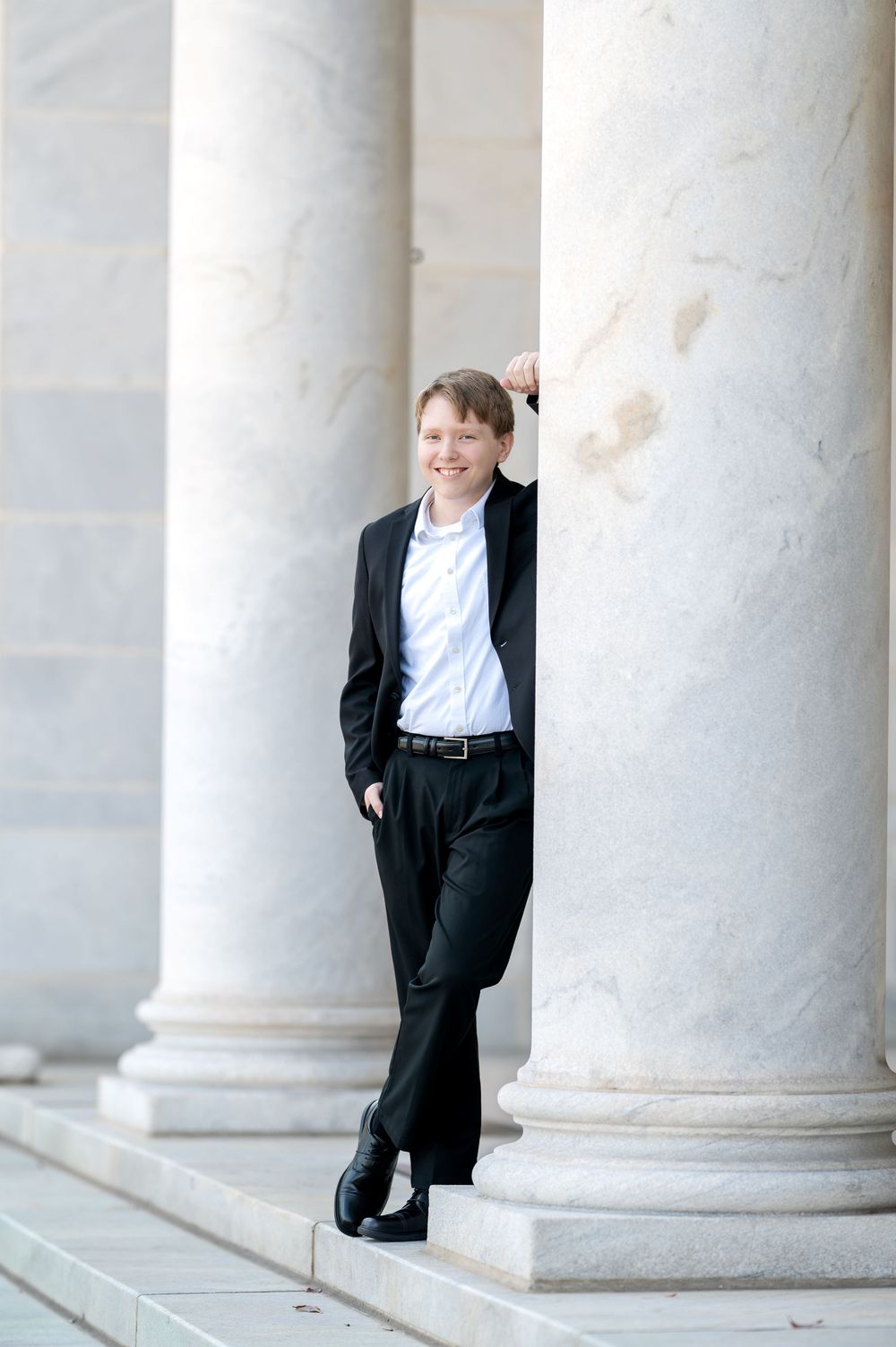 Smiling young man in black suit leaning against marble column on steps.