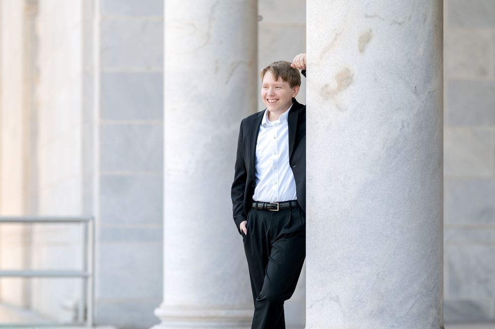 Young man in black blazer leaning against marble column, smiling outdoors.