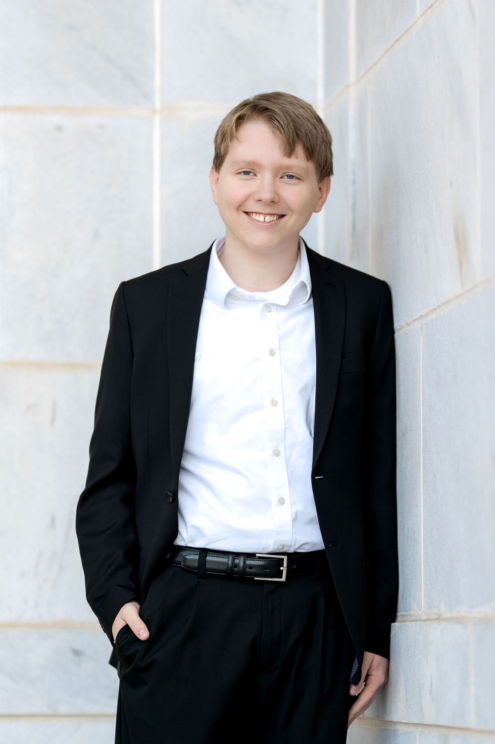 Smiling teen boy in black blazer and white shirt leaning against marble wall.