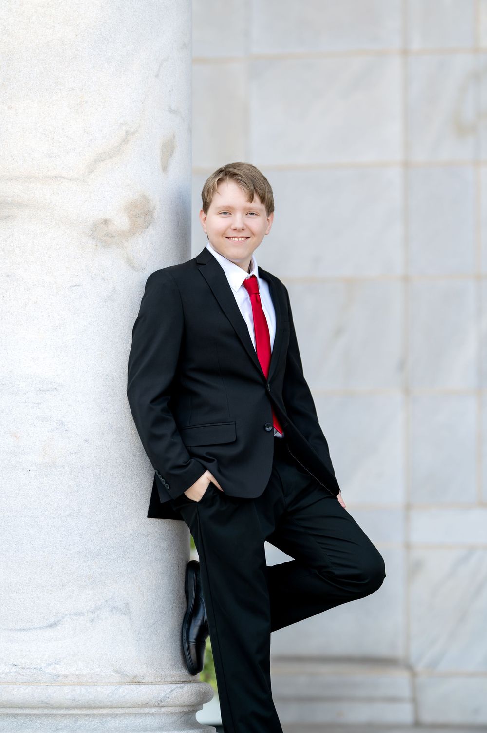 Young boy in black suit and red tie leaning against marble column, smiling.