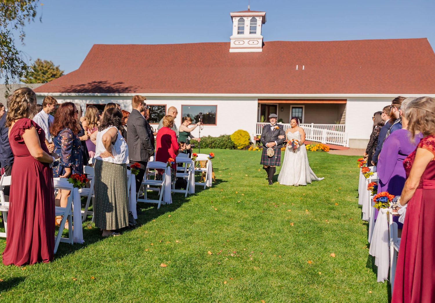 A panoramic view of wedding guests gathered on a green lawn outside a white barn on a sunny day.