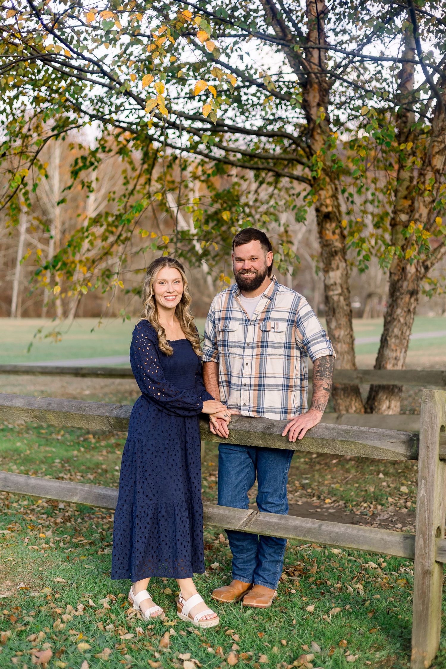 A couple shares a romantic autumn moment together by a wooden fence surrounded by fall foliage and trees.