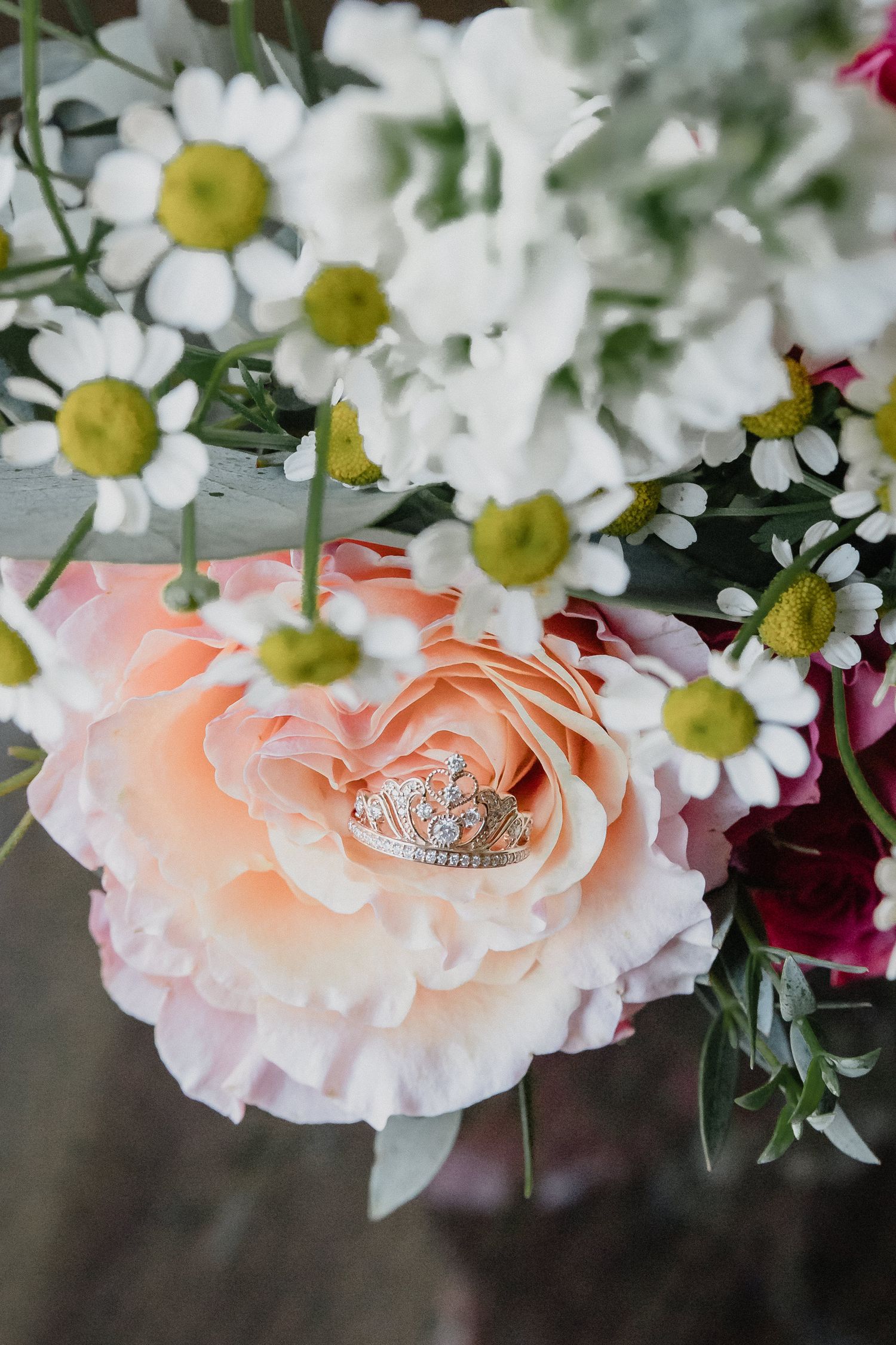 Wedding rings nestled in a delicate peach-colored rose bloom surrounded by white daisies and burgundy flowers.