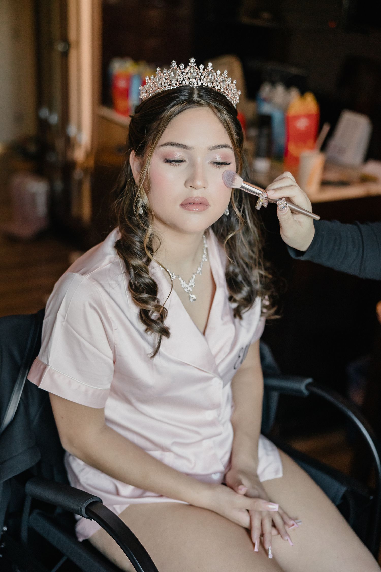 A person wearing a tiara and pink satin robe sits in a dimly lit room during bridal preparation.