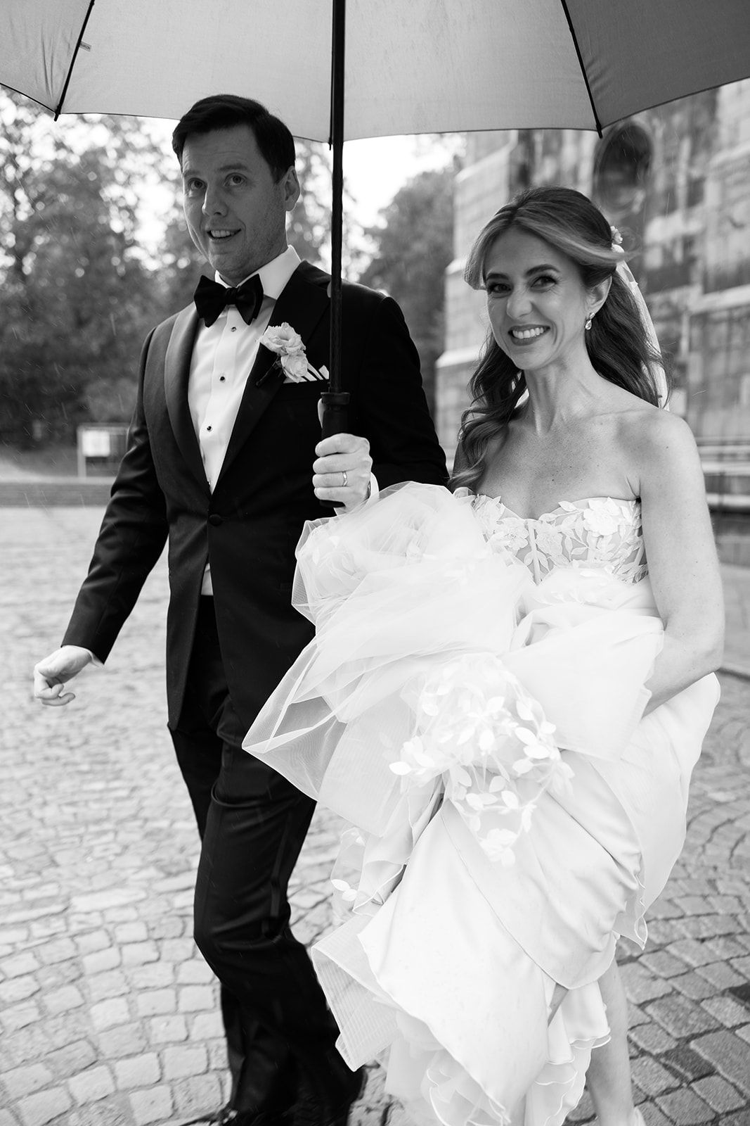 A bride in a white strapless gown and groom in a tuxedo share an umbrella on a rainy cobblestone street.
