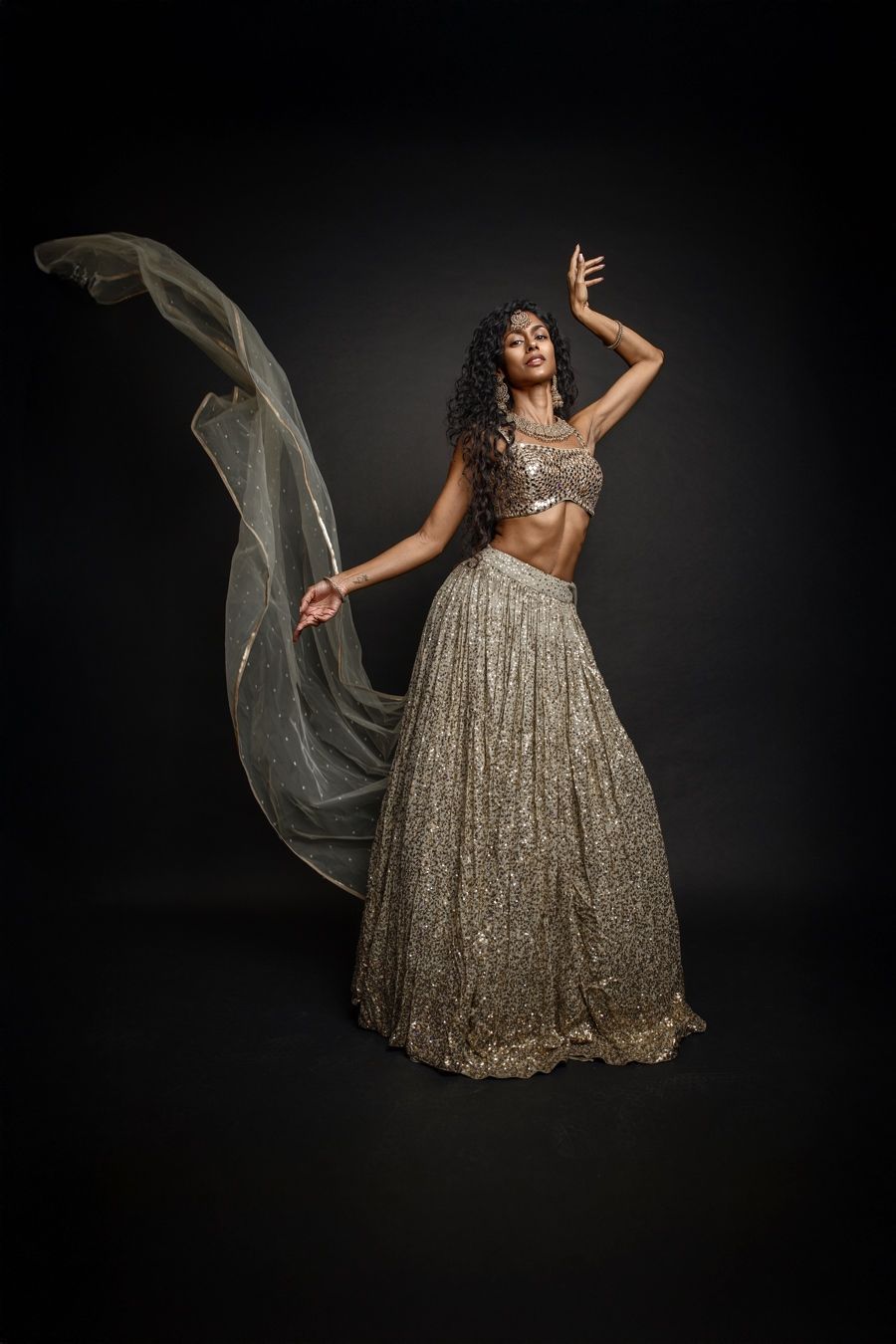 A model in a sparkling gold ballgown poses dramatically against a dark background with flowing fabric in motion.