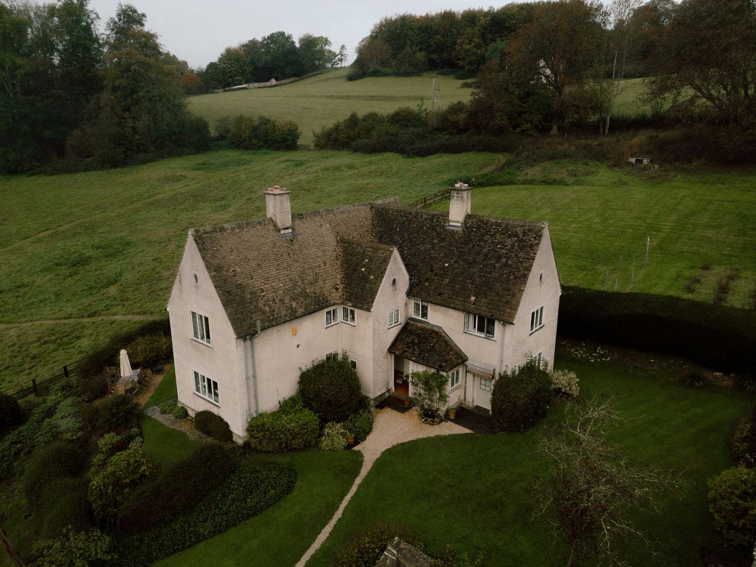 A charming Cotswold stone cottage with gabled roof sits nestled among rolling green hills and countryside gardens.