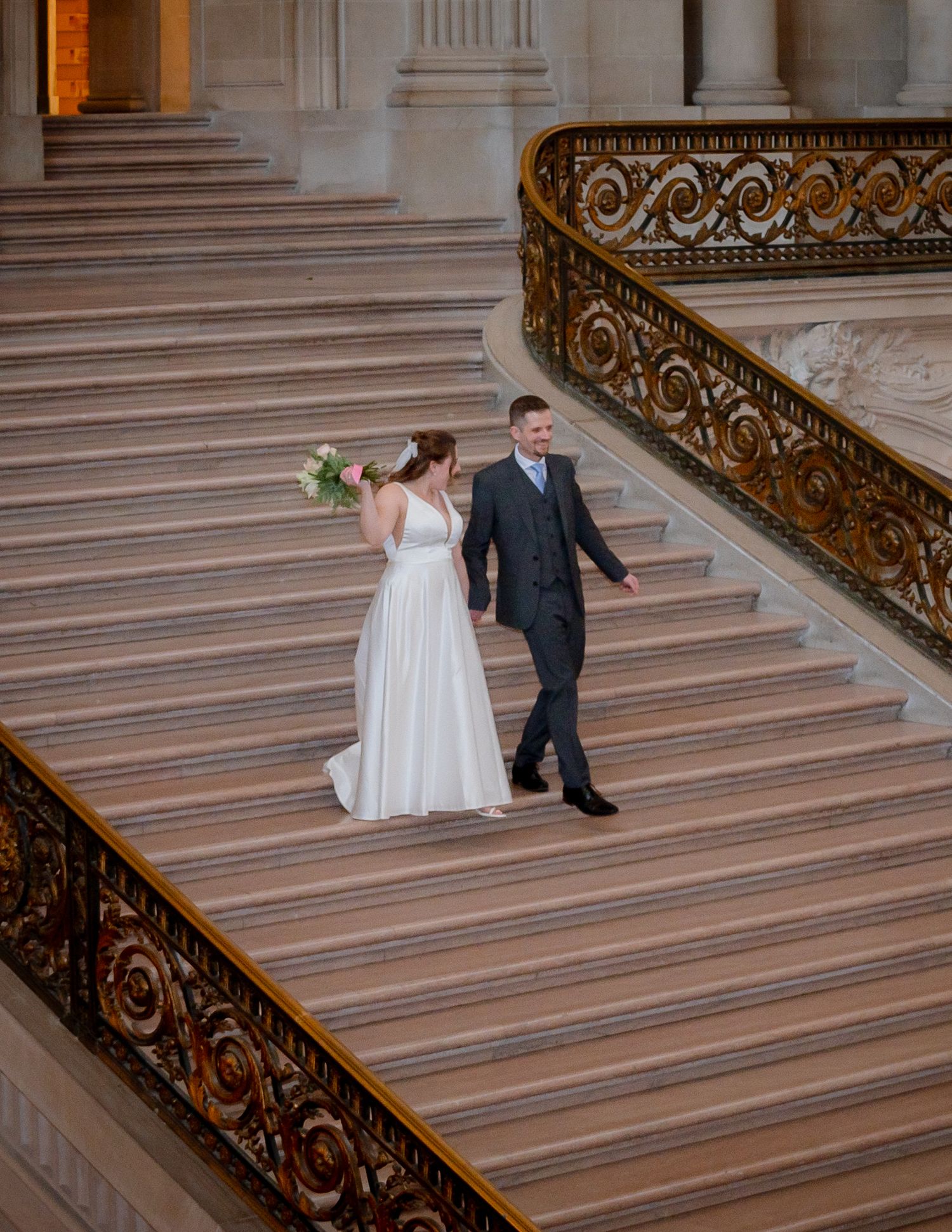 A wedding couple stands on the grand marble staircase of a historic building.
