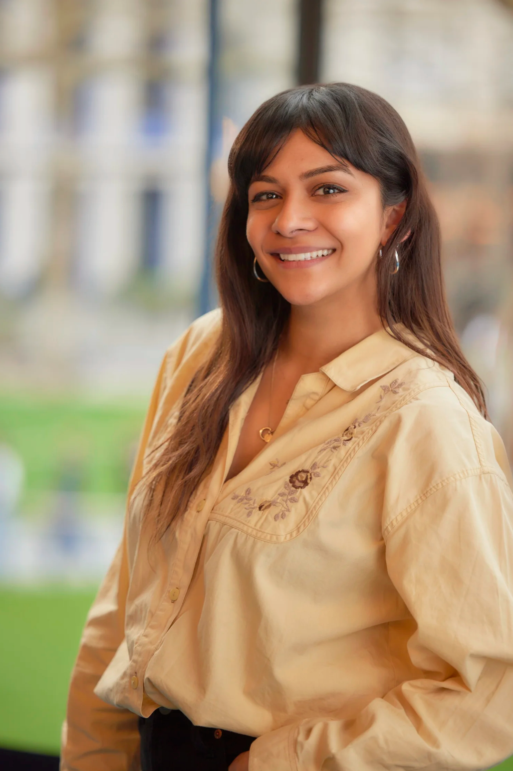 Professional portrait of person in beige embroidered blouse against blurred background.