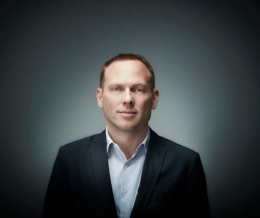 Professional headshot of someone in a dark suit jacket and light shirt against a gradient background.