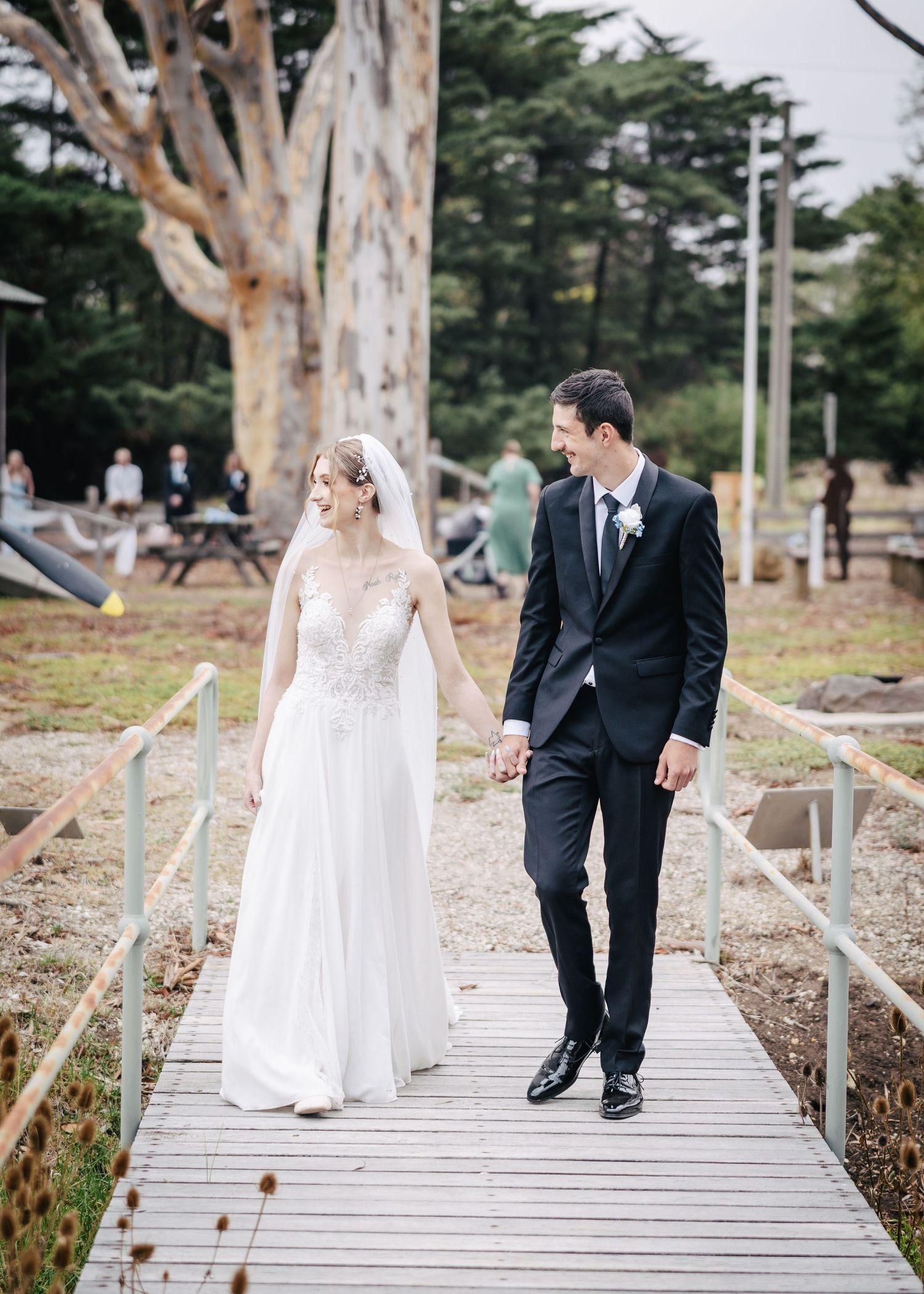 Wedding couple walks together on wooden boardwalk surrounded by trees in romantic outdoor setting.