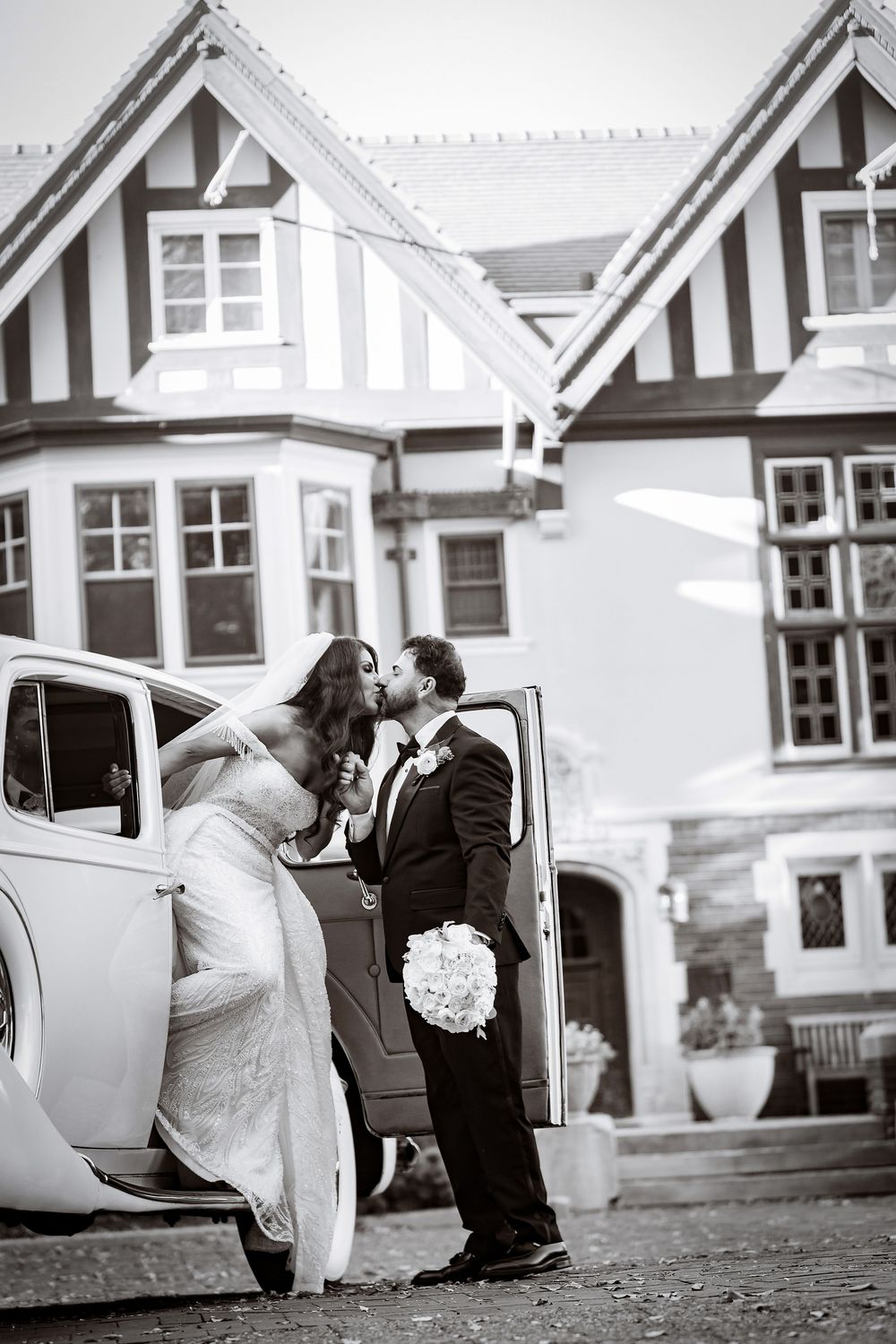 Black and white wedding photo sequence showing couple in formal attire by vintage car outside Tudor-style building.