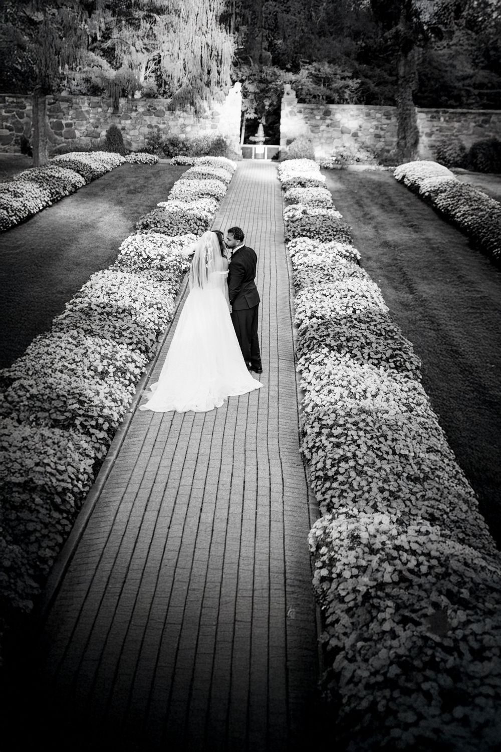 A romantic black and white wedding photograph on a garden path lined with blooming flowers leading to a stone archway.