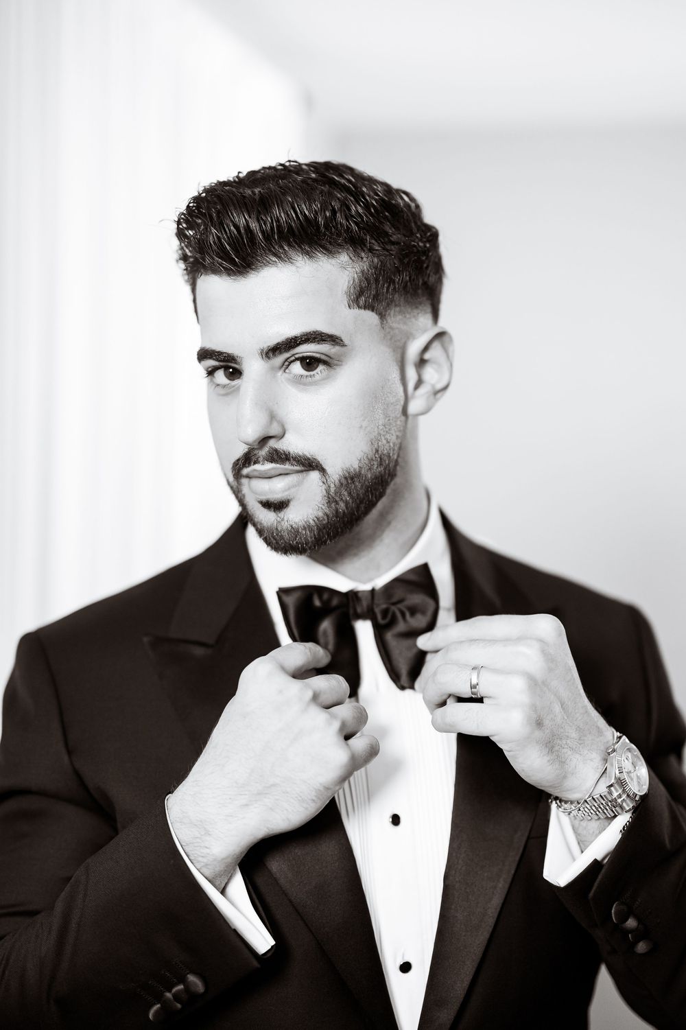 A well-dressed groom in a black tuxedo adjusting his bow tie, black and white portrait photo.