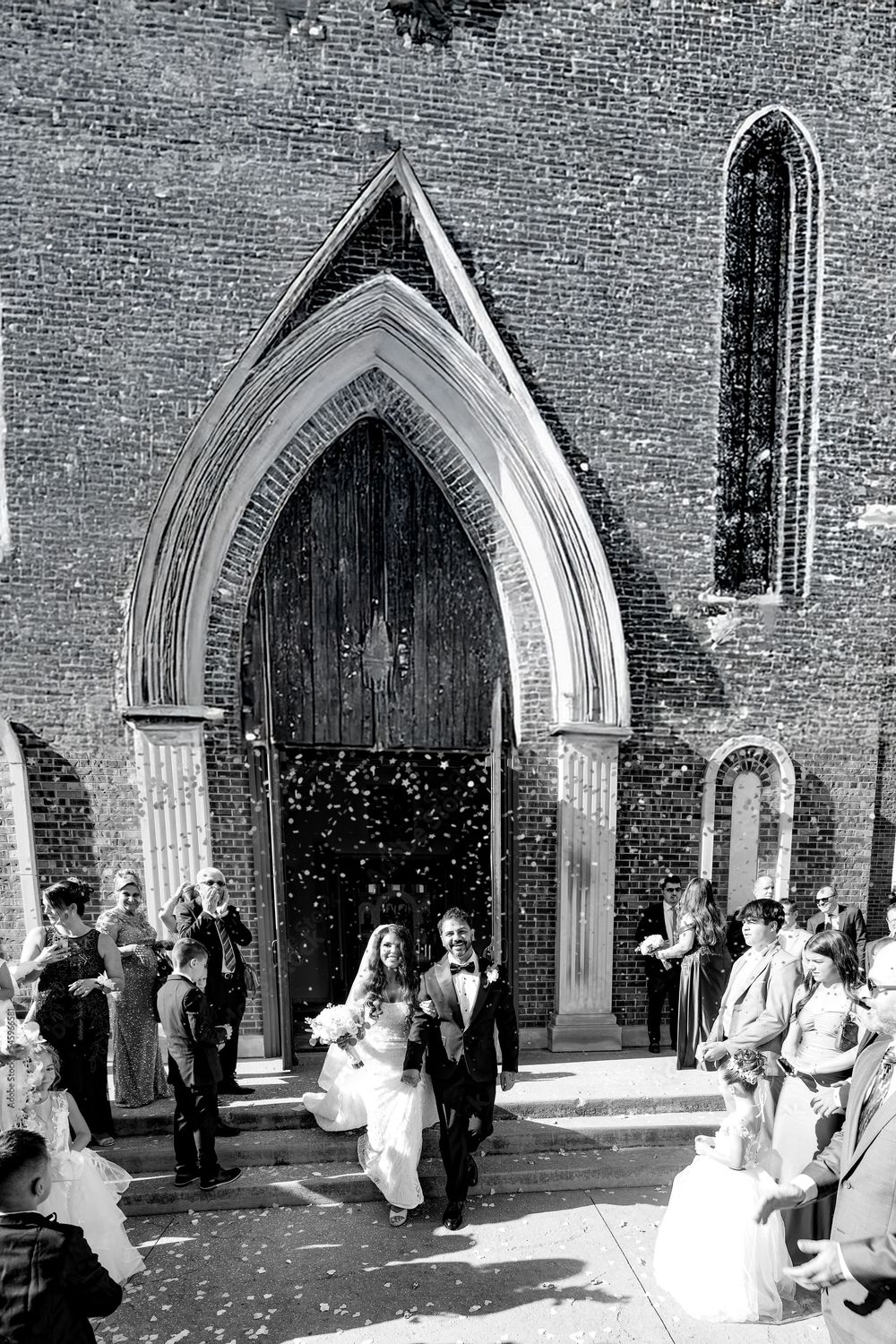 Wedding guests celebrate outside a historic brick church with Gothic arched doorways during a confetti toss ceremony.