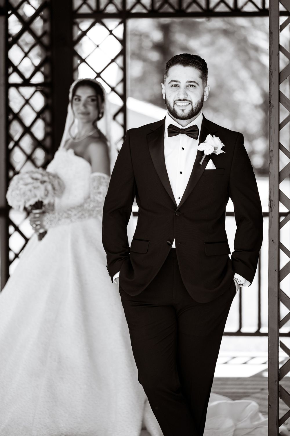 Black and white wedding photo of smiling groom in tuxedo with bride in ball gown holding bouquet in background.