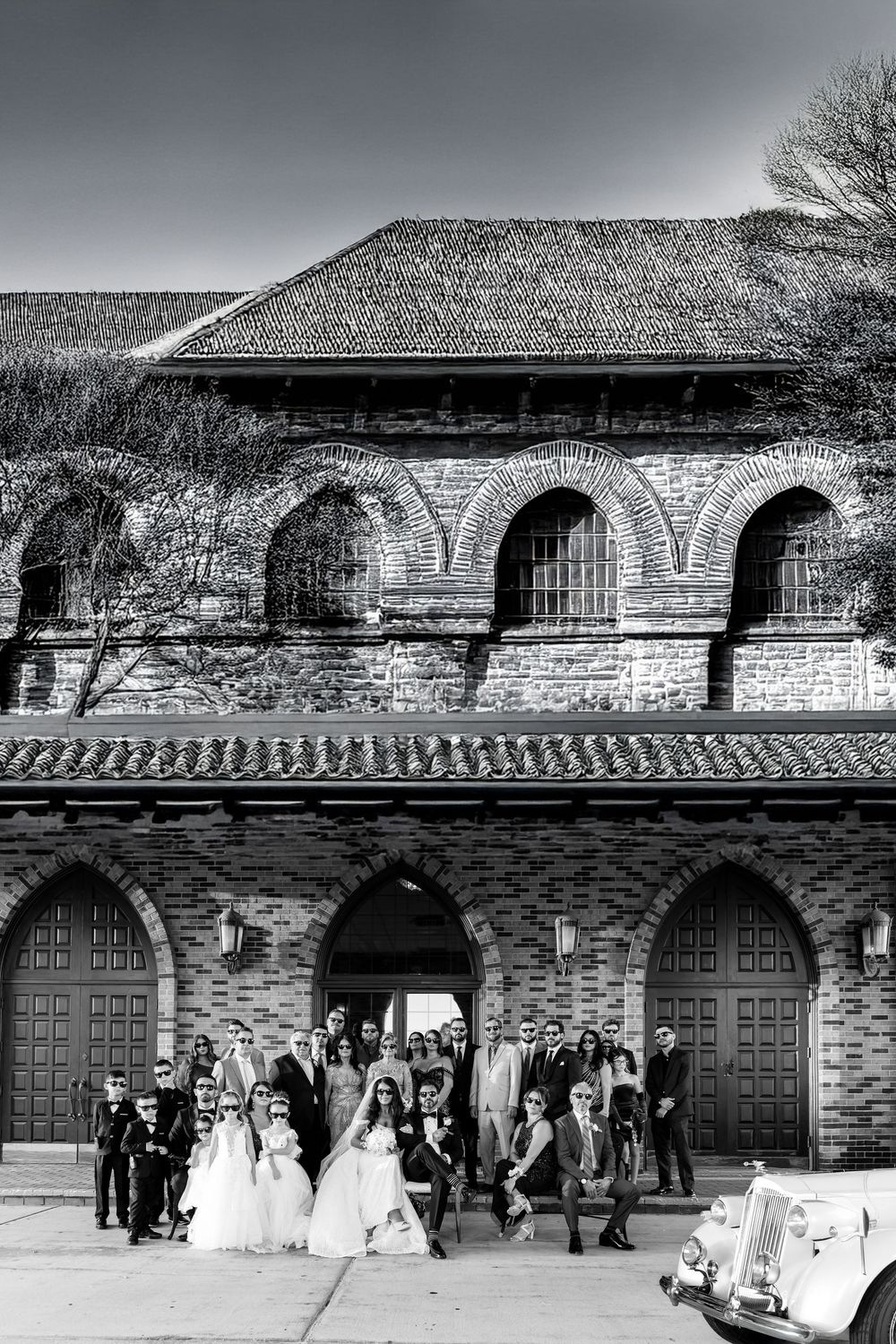 A panoramic black and white photo of a stone building facade with arched doorways and a wedding party gathering outside.