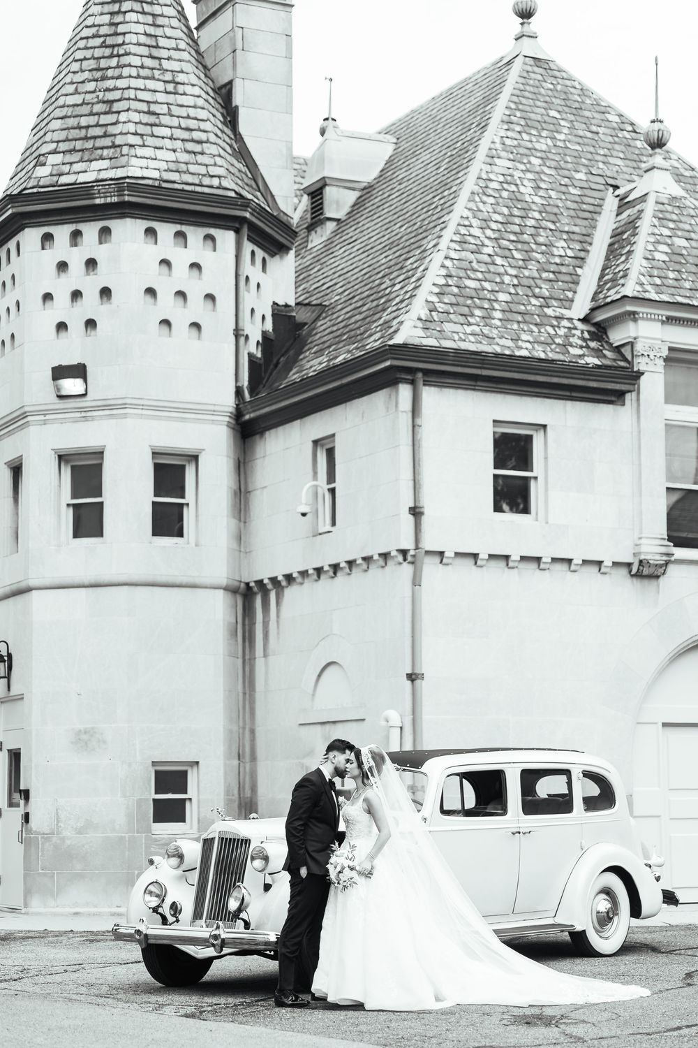 Black and white photo of bride and groom kissing beside a vintage car in front of a castle-like building.