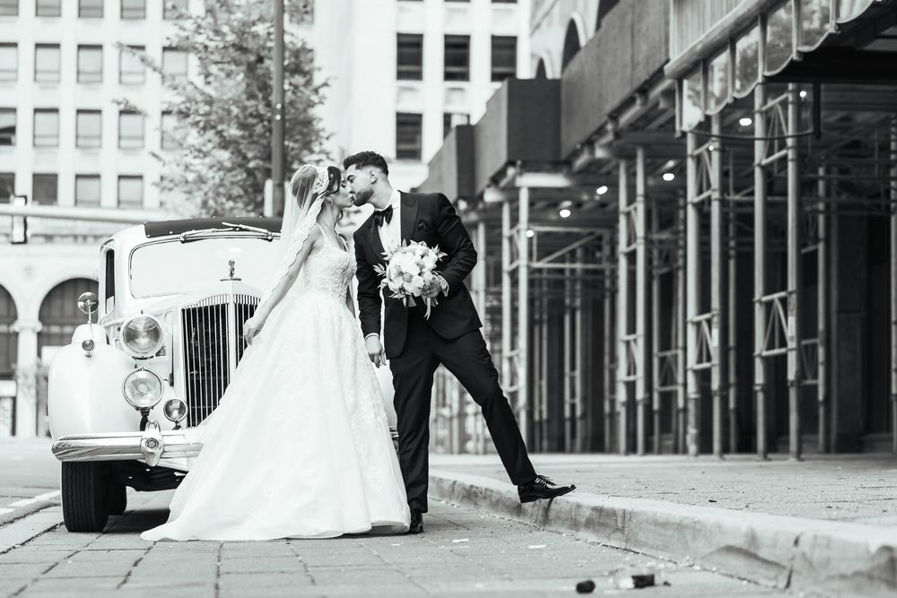 Black and white photo of a bride and groom kissing beside a vintage car on a city street.