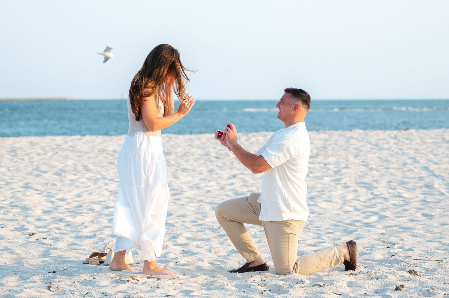 A couple in white outfits shares a romantic proposal moment on a pristine white sandy beach at sunset.