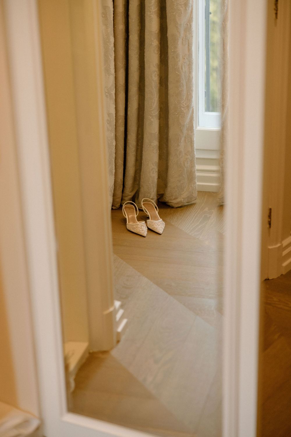 A pair of elegant white bridal shoes sits on a light wooden floor next to beige curtains reflected in a mirror.