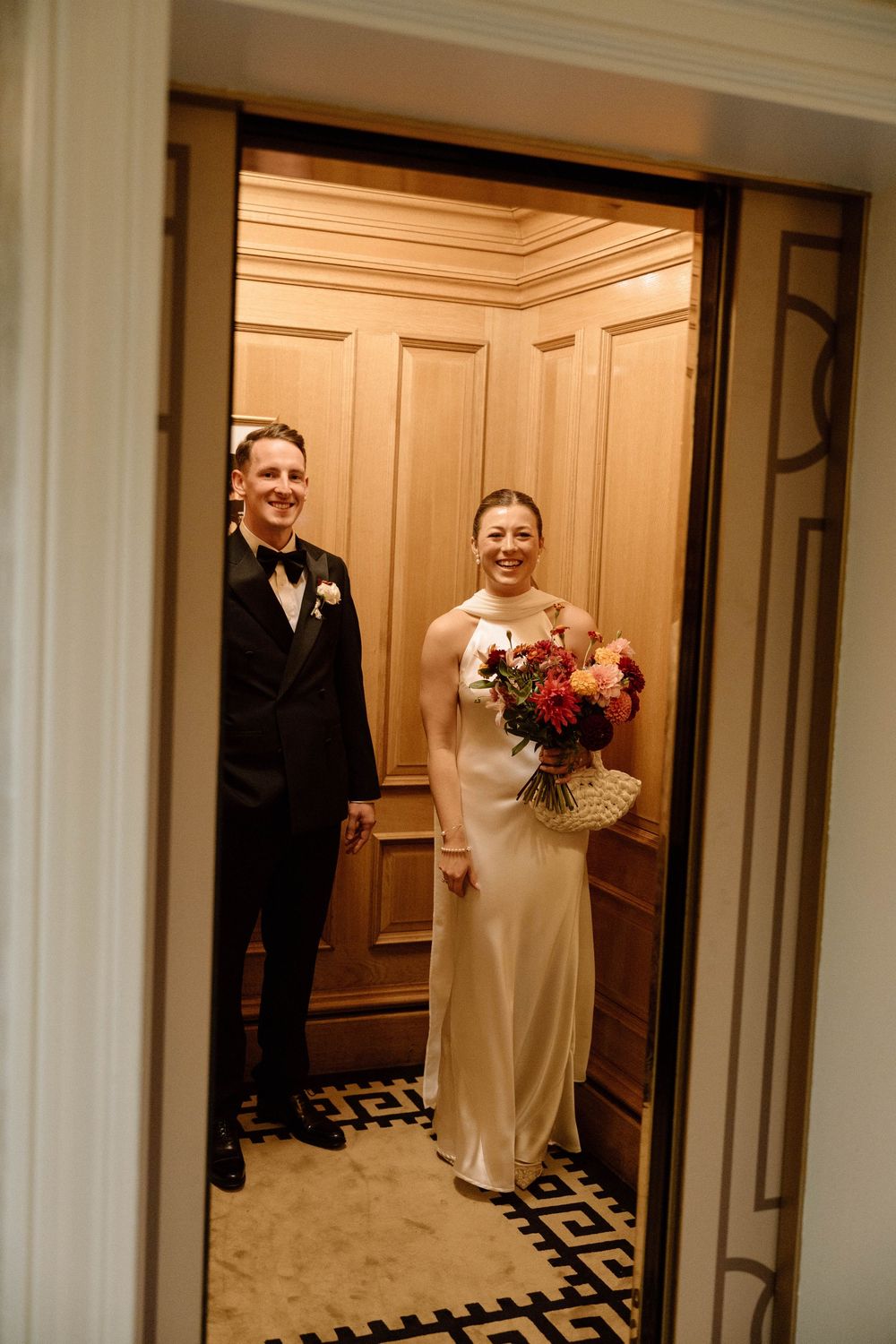 Bride and groom share playful moments in hotel hallway with geometric patterned carpet and cream walls.
