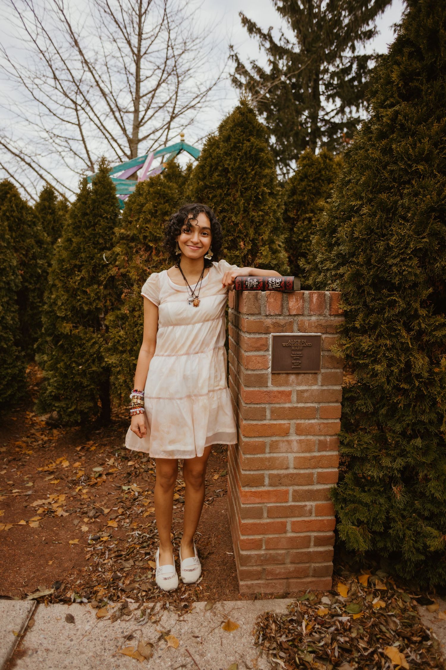 A casual outdoor photo series showing someone in a light-colored dress posing by evergreen trees and a brick pillar.
