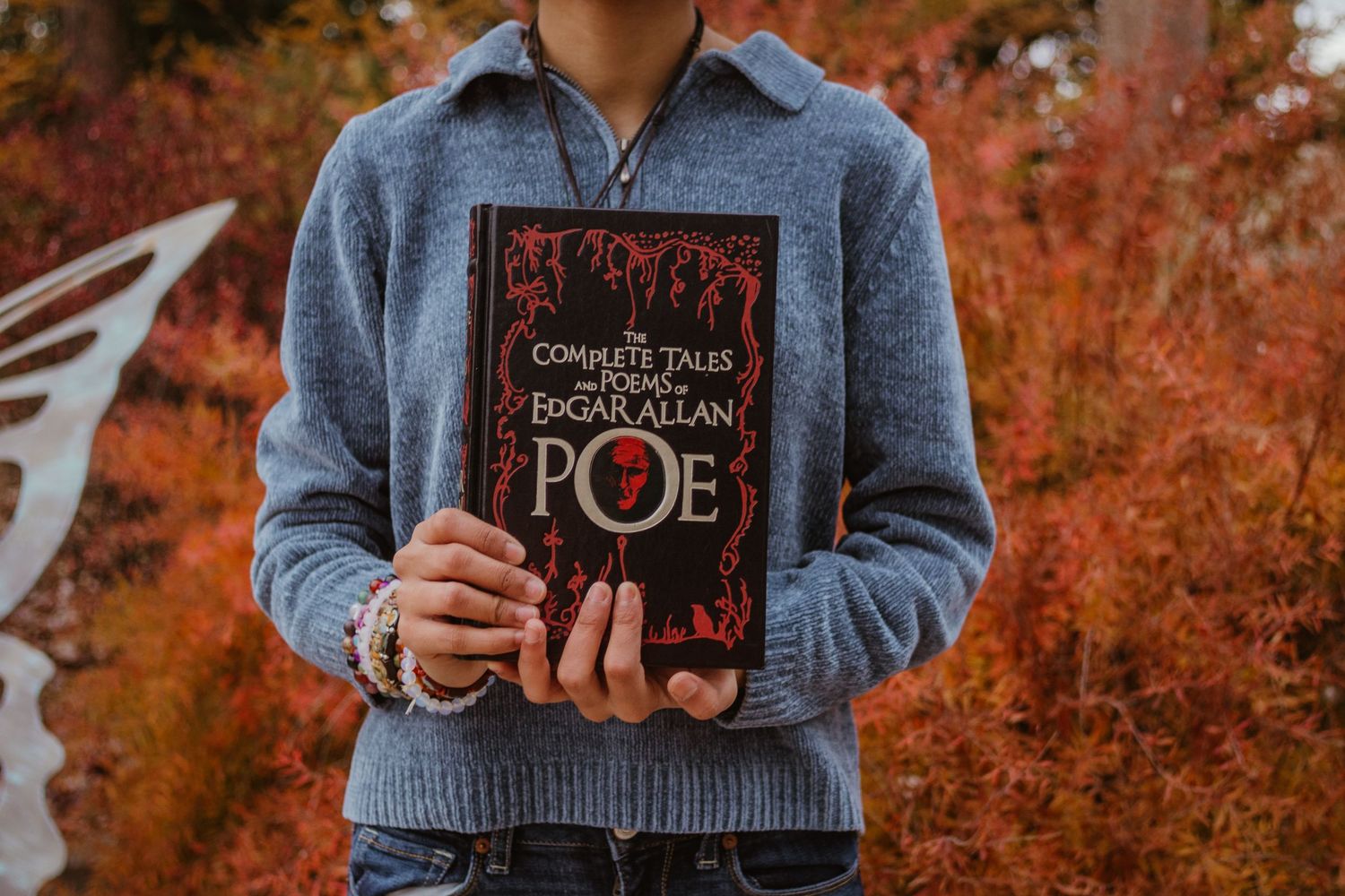 Someone holding and displaying Edgar Allan Poe books against a backdrop of vibrant fall foliage.