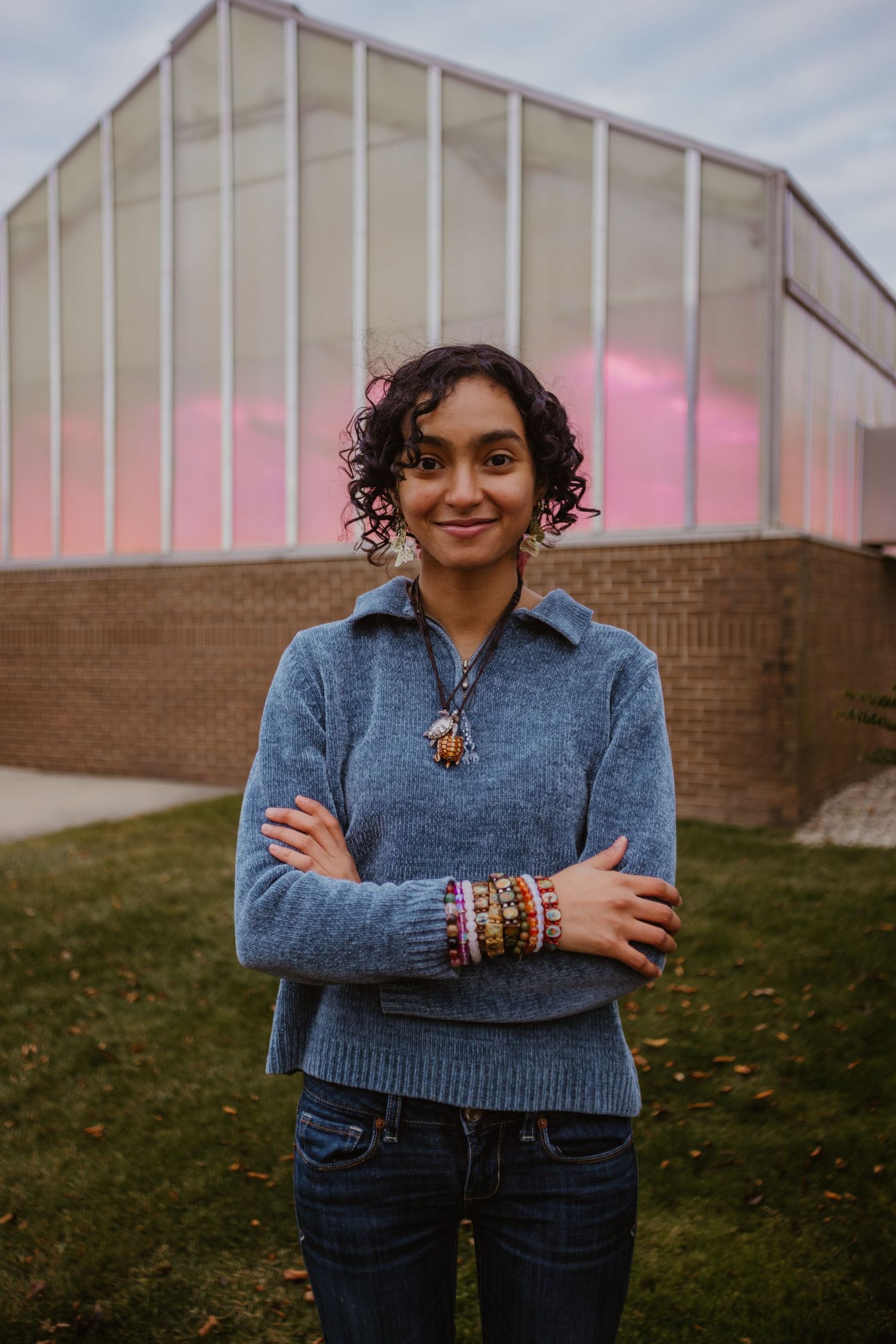 A person wearing a blue sweater and jeans poses against a brick building with glowing pink windows.