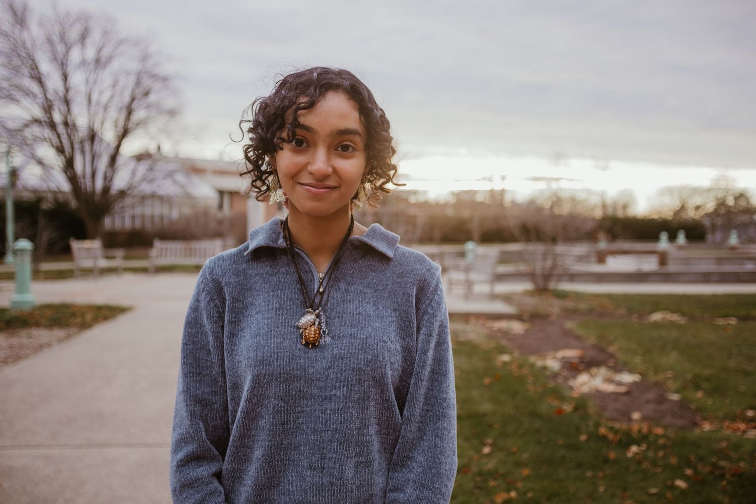 A person wearing a blue sweater stands outdoors in autumn with trees and buildings in the background.